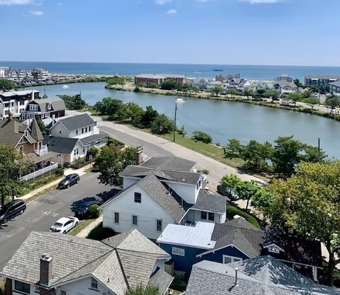 A scenic view of a coastal neighborhood with houses, a road, trees, a body of water, and the ocean in the background under a clear blue sky.
