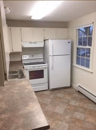 A small kitchen with white cabinets, a white refrigerator, a white electric stove with oven, a double stainless steel sink, and a window showing an outdoor view. The floor has a checkered tile pattern in neutral tones.