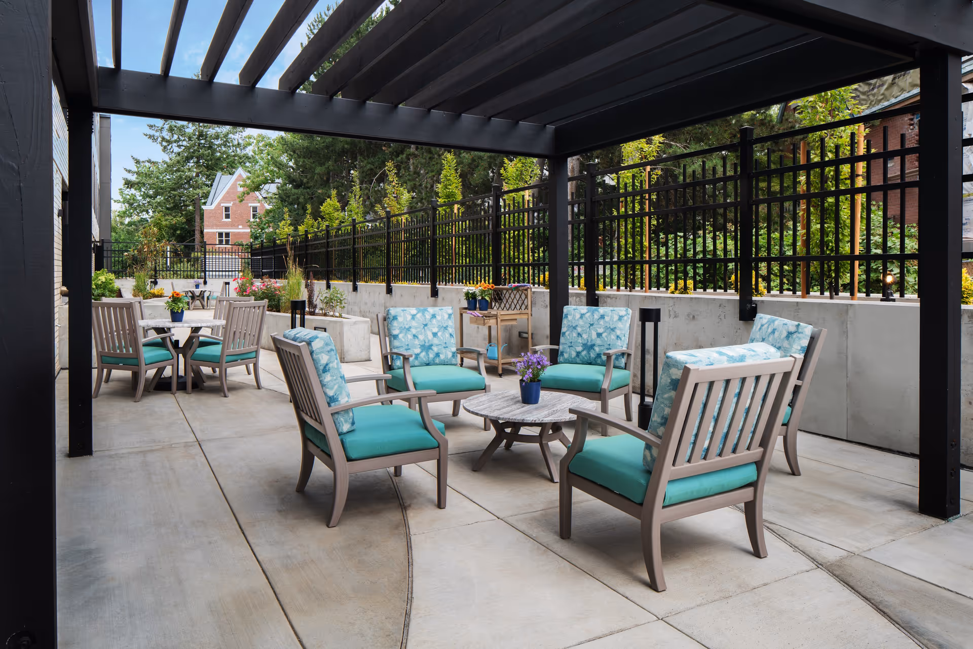 Outdoor patio area with cushioned chairs and small tables under a pergola, surrounded by a black metal fence and greenery, with a brick building visible in the background.