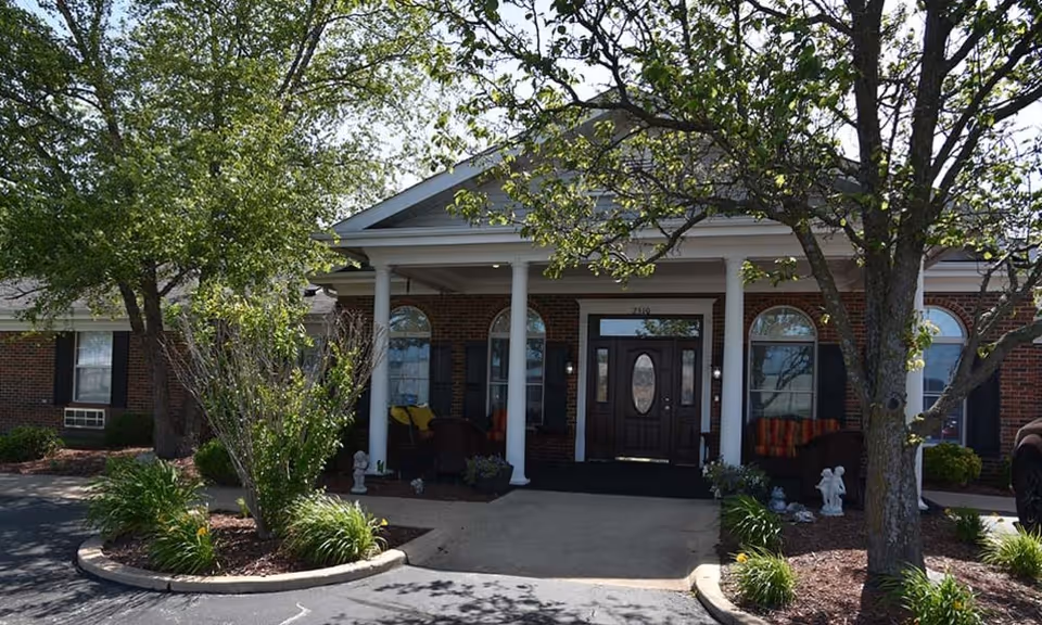 Front entrance of a brick assisted living building with white columns, a covered porch, seating, and trees in the foreground.