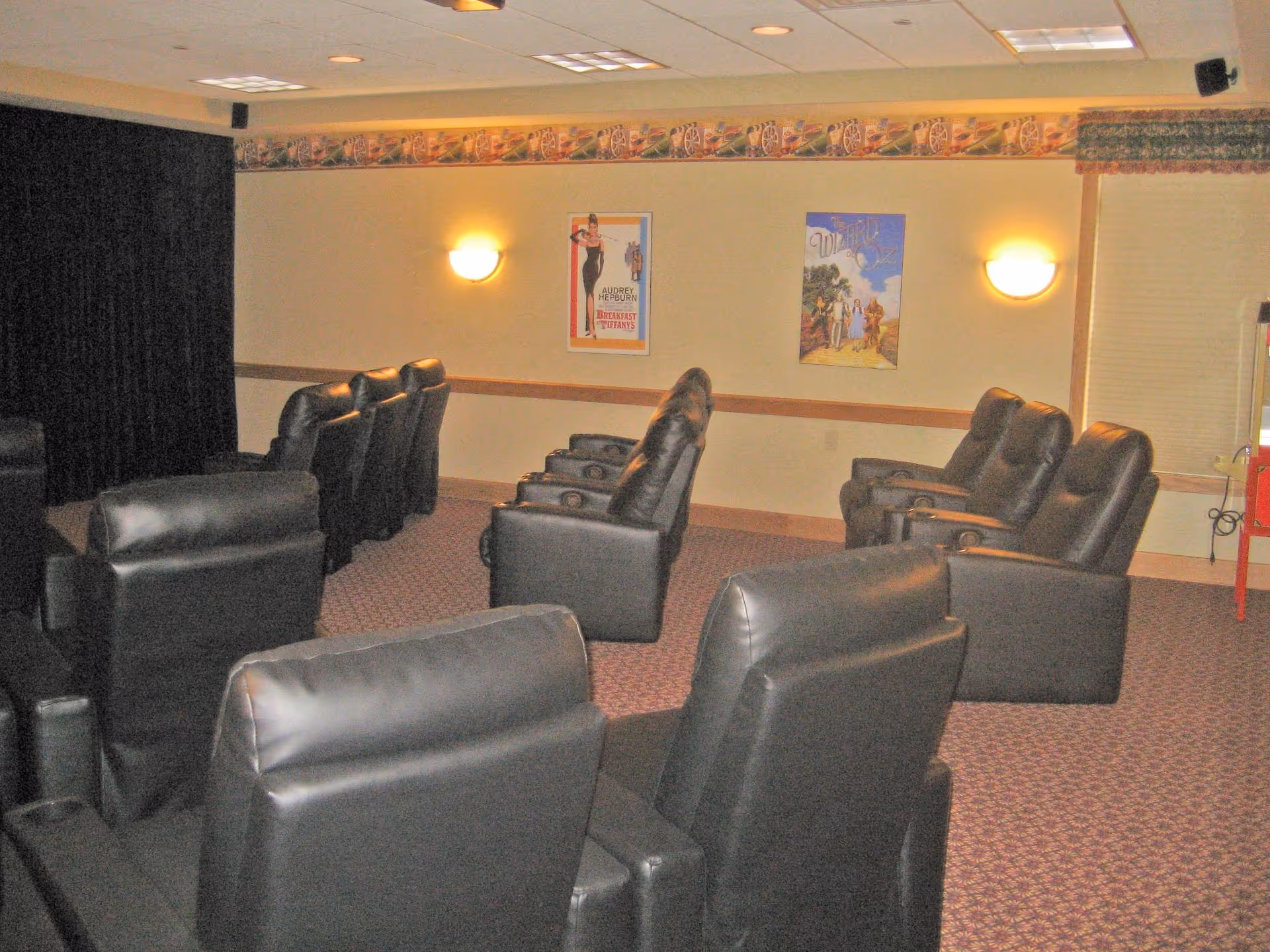 A small theater room with multiple rows of black leather recliner chairs facing a black curtain, with two movie posters on the beige wall and wall-mounted lights illuminating the space.