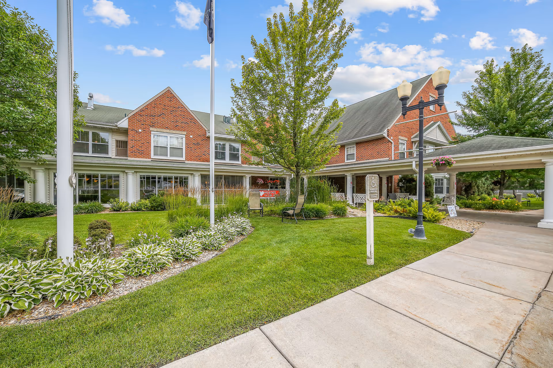 Exterior view of Heathers Manor, a two-story brick building with white trim, surrounded by green grass, trees, and landscaped plants. A covered driveway entrance is visible on the right side, along with a sidewalk and a lamppost. The sky is partly cloudy.