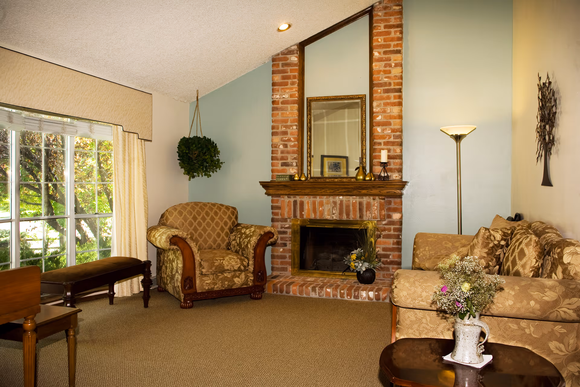 A cozy living room with a brick fireplace centered on a light blue wall. Above the fireplace is a large mirror and decorative items on the mantel. To the left is a patterned armchair with wooden armrests, next to a large window with cream-colored curtains. To the right is a floral-patterned sofa with a floor lamp behind it and a small wooden coffee table in front holding a vase of flowers. A hanging plant is suspended near the armchair, and a decorative wall piece is mounted on the right wall.