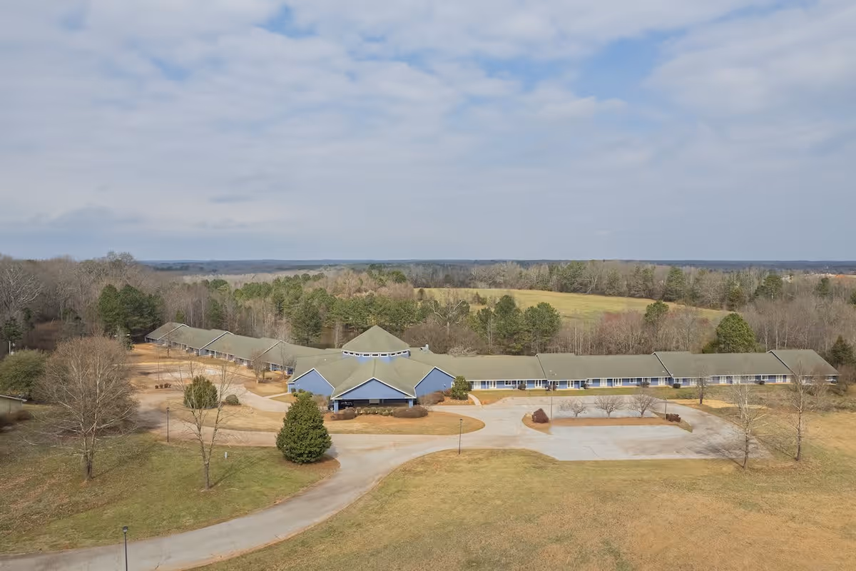 Aerial view of The Overlook Senior Living facility showing a long, single-story building with a green roof surrounded by trees and open grassy areas under a partly cloudy sky.