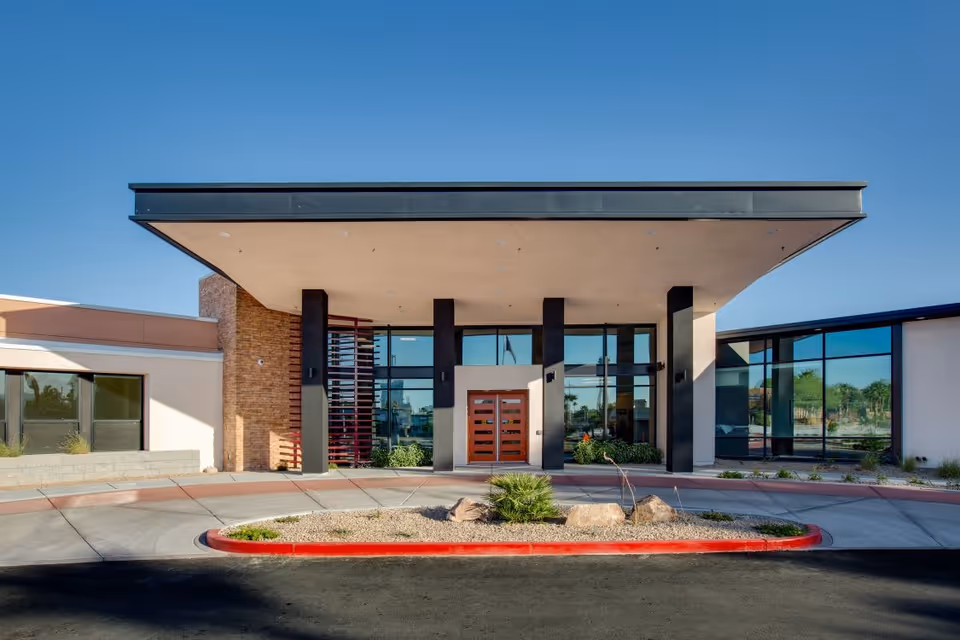 Front exterior view of Lumina Las Vegas facility with a modern entrance featuring large glass windows, a covered entryway supported by black pillars, and a landscaped area with rocks and plants in front.