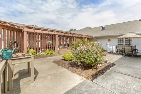 Outdoor courtyard area with a concrete walkway, a garden bed with flowering bushes, a wooden pergola with hanging decorations, a small table with gardening supplies, and a patio table with chairs and an umbrella next to a white building.