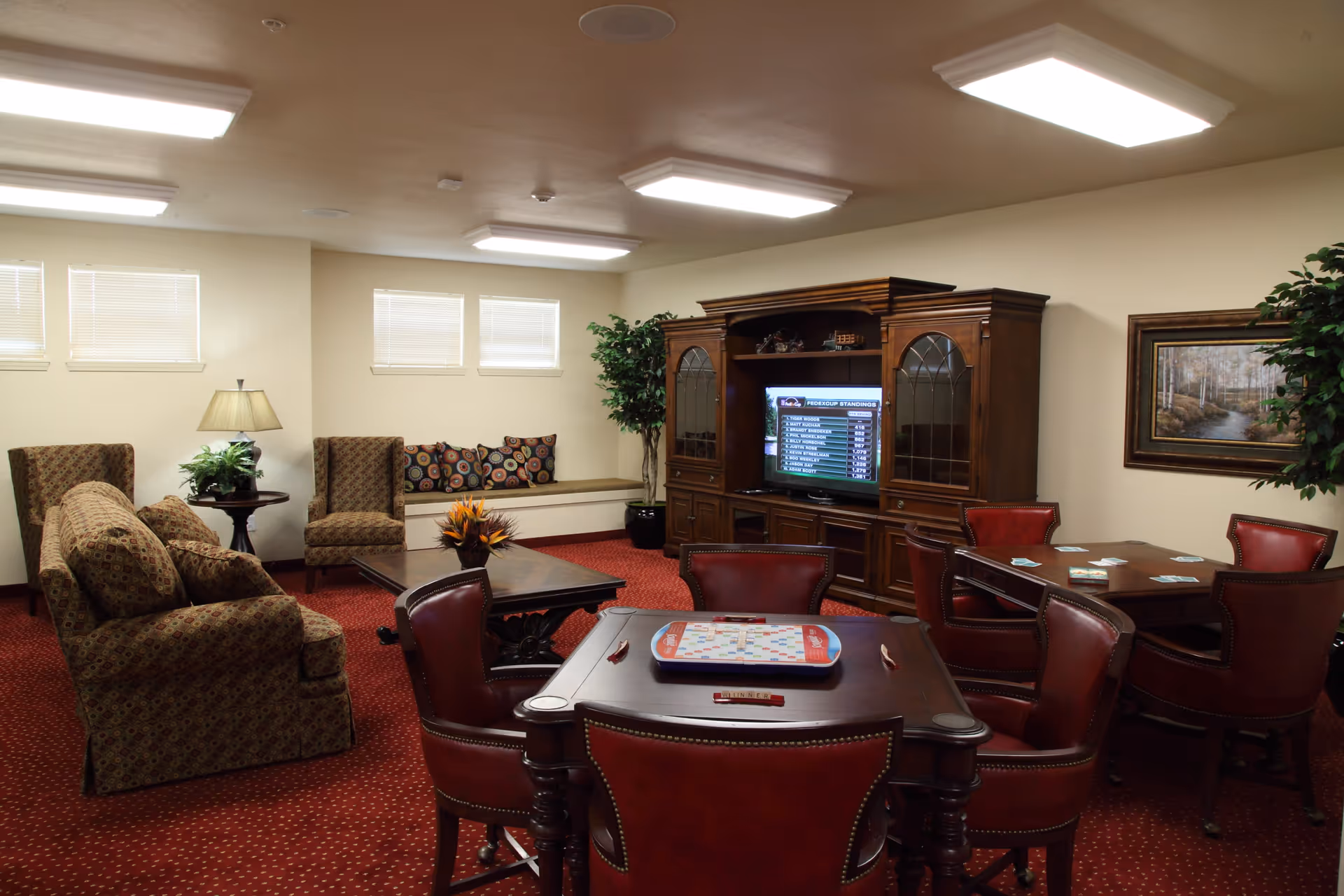 A cozy common room with patterned upholstered chairs and sofas, a wooden coffee table with a flower arrangement, two tables with red leather chairs set up for board games, a large wooden entertainment center with a TV, and decorative plants and artwork on the walls.