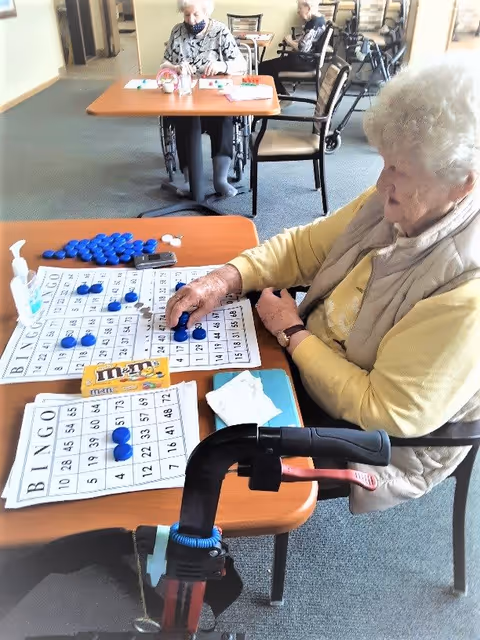 An elderly woman playing bingo at a table in a senior living facility. She is placing blue markers on a bingo card. On the table are additional bingo cards, a box of M&M's, and a bottle of hand sanitizer. In the background, two other elderly individuals are seated at another table, one in a wheelchair. The setting appears to be a common activity area with chairs and tables.
