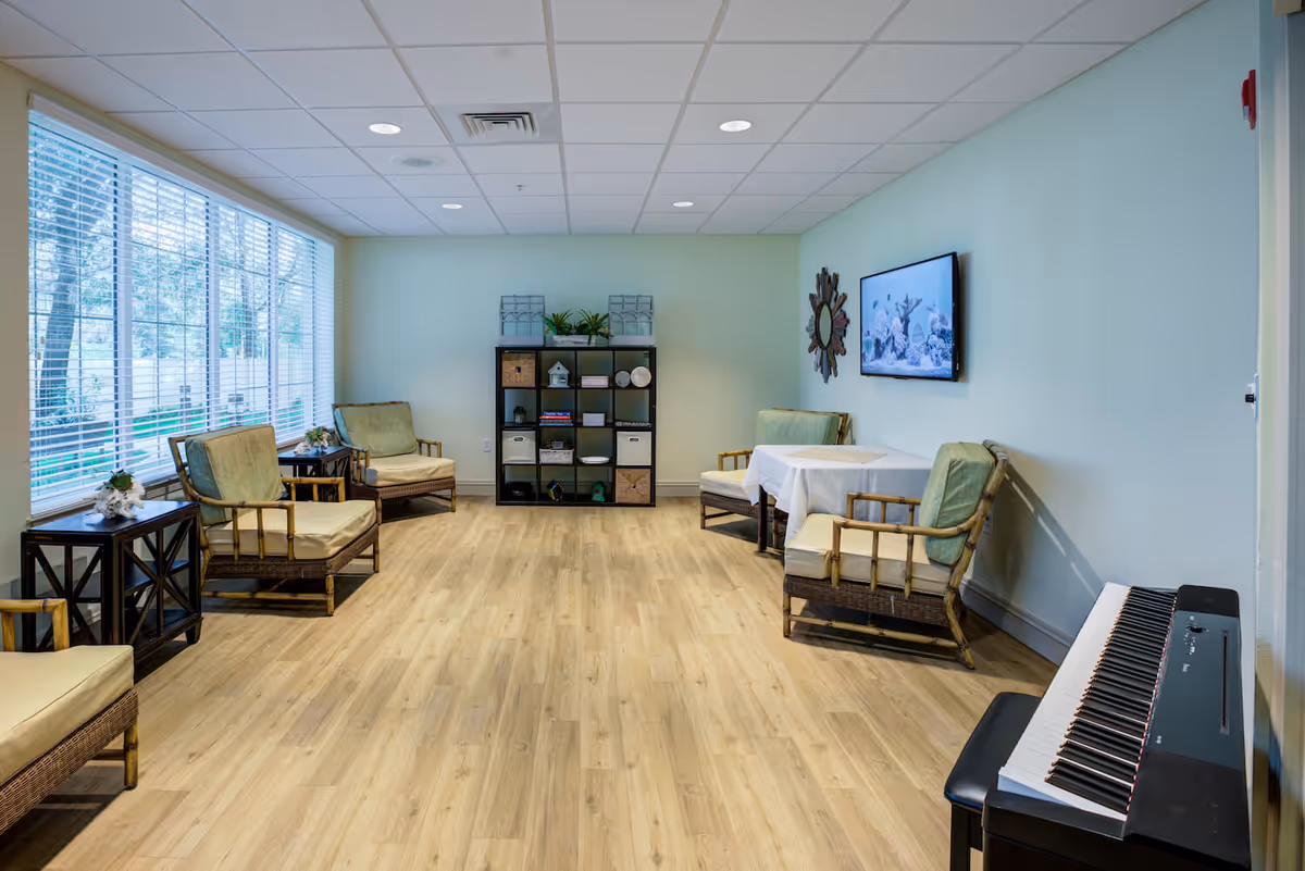 A bright and spacious common area with light wood flooring, several cushioned chairs arranged along the walls, a small table covered with a white cloth, a black shelving unit with decorative items, a wall-mounted TV displaying an underwater scene, and a digital piano with a bench in the foreground. Large windows with blinds allow natural light to fill the room.