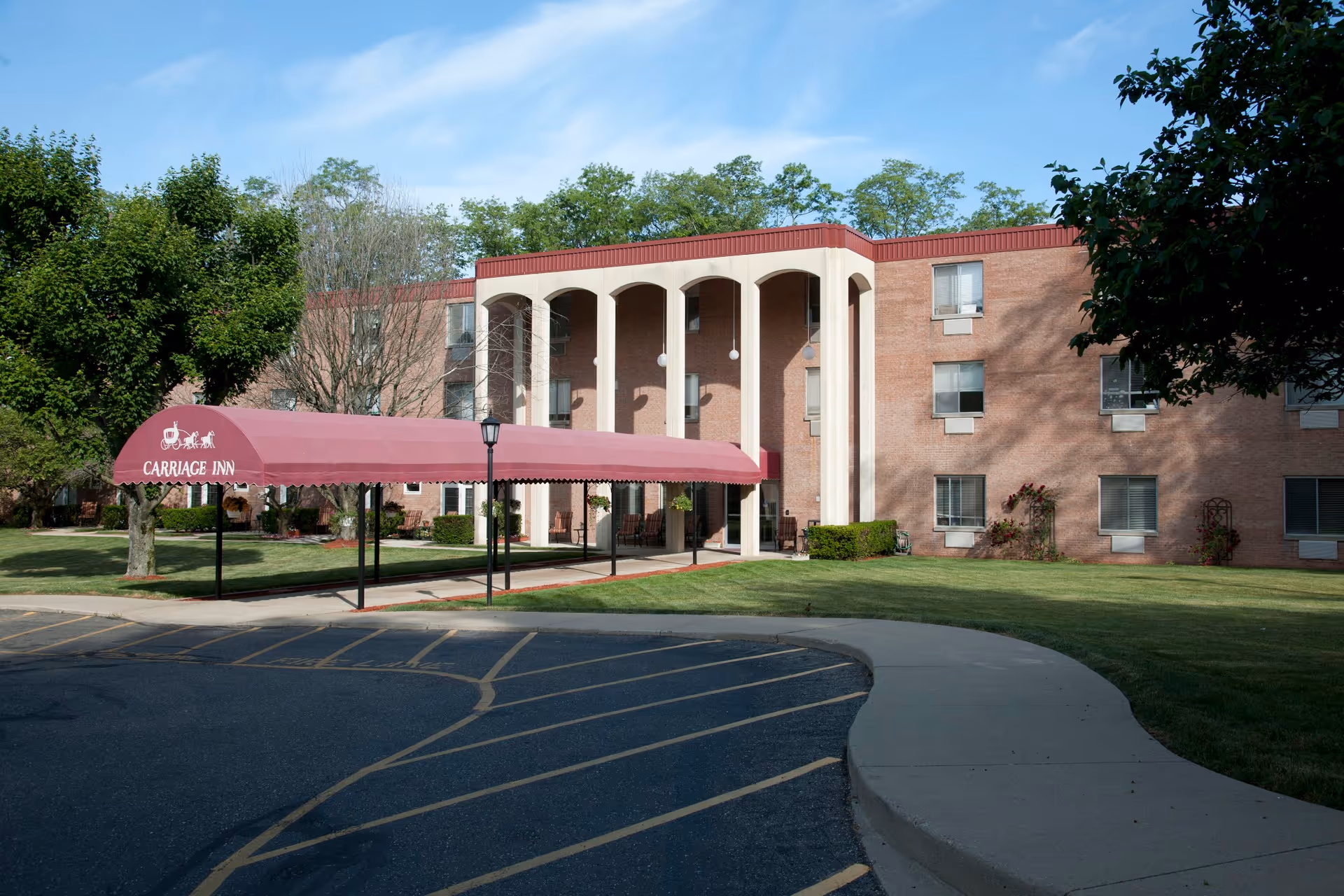 Exterior view of a three-story brick building with a covered entrance canopy labeled 'Carriage Inn'. The building is surrounded by green grass, trees, and a paved driveway with parking spaces.