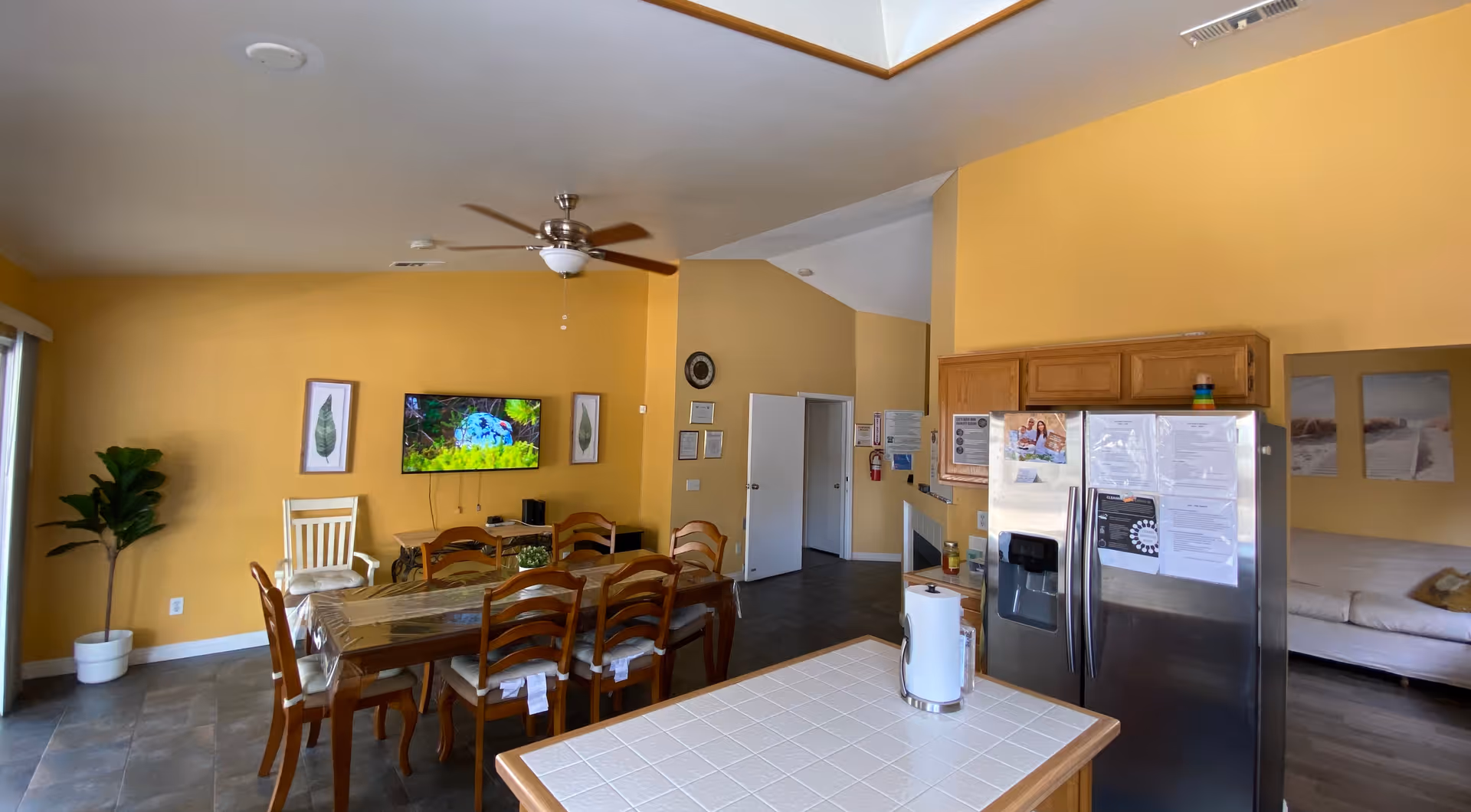 Interior view of a senior care facility dining and living area with yellow walls. There is a wooden dining table with eight chairs, a ceiling fan, a wall-mounted TV displaying a nature scene, and framed leaf artwork on the wall. To the right, there is a kitchen area with a stainless steel refrigerator and wooden cabinets. A white tiled kitchen island with a paper towel holder is in the foreground. In the background, there is a white couch and two framed pictures on the wall.