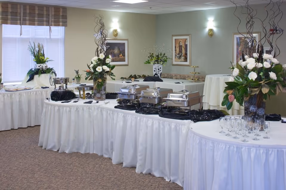 A buffet setup in a dining area with tables covered in white tablecloths, chafing dishes, floral centerpieces, and glassware. The room has beige walls with framed artwork and wall sconces, and a window with a valance.