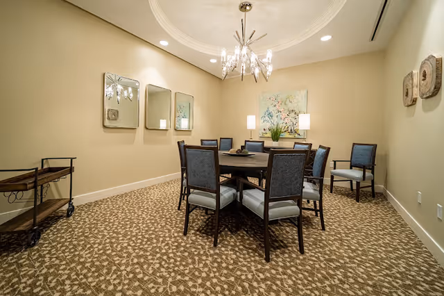 A small dining room with a round wooden table surrounded by upholstered chairs, a chandelier overhead, patterned carpet, and wall art.