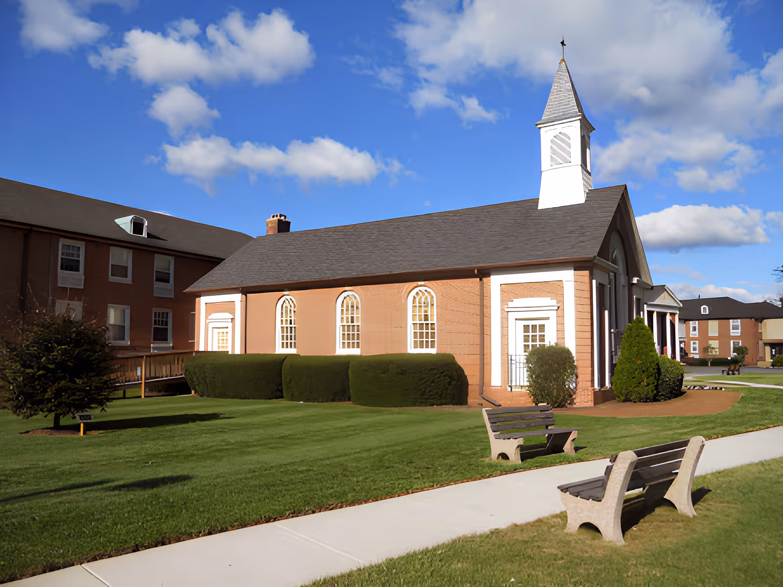 Exterior view of a brick building with white trim and a steeple, surrounded by green grass, bushes, and a sidewalk with benches under a blue sky with scattered clouds.