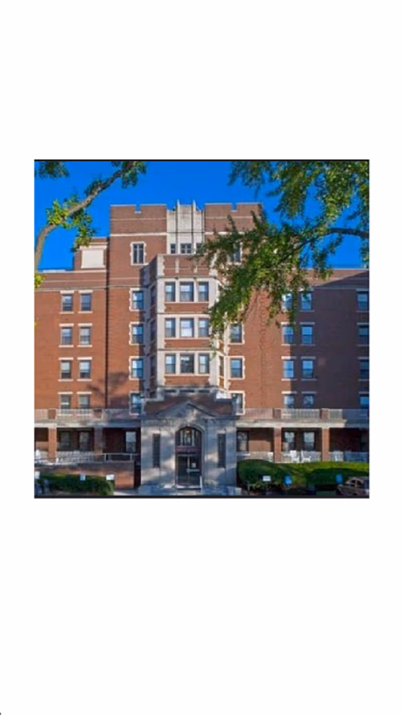 Front exterior view of a multi-story brick building with multiple windows and a central entrance, partially framed by tree branches with green leaves against a clear blue sky.