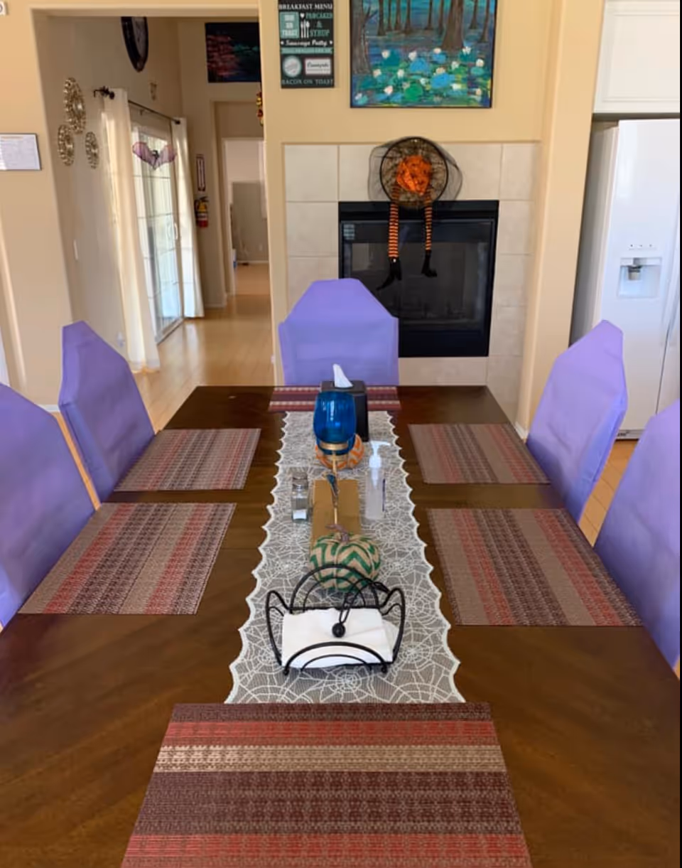 A dining room with a wooden table set with six purple-covered chairs. The table has a lace runner with a blue candle holder, a tissue holder, a small decorative pumpkin, a salt shaker, and a bottle of hand sanitizer. In the background, there is a fireplace decorated with a Halloween witch hat and legs, a painting above it, and a white refrigerator to the right.