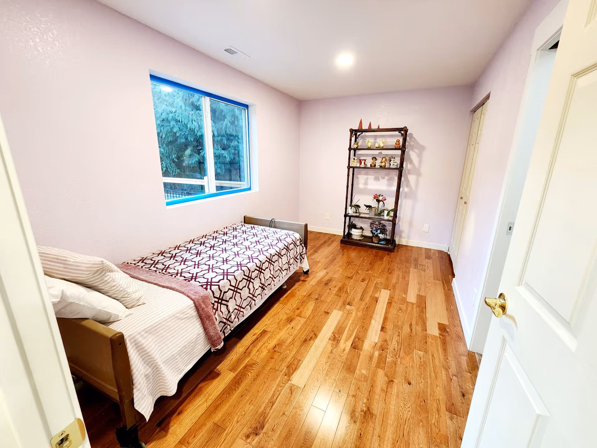 A small bedroom with light purple walls and wooden flooring. There is a single bed with a patterned blanket and pillows on the left side under a window showing greenery outside. On the far wall, there is a dark wooden shelving unit with decorative items and plants. A white door is partially open on the right side of the image.