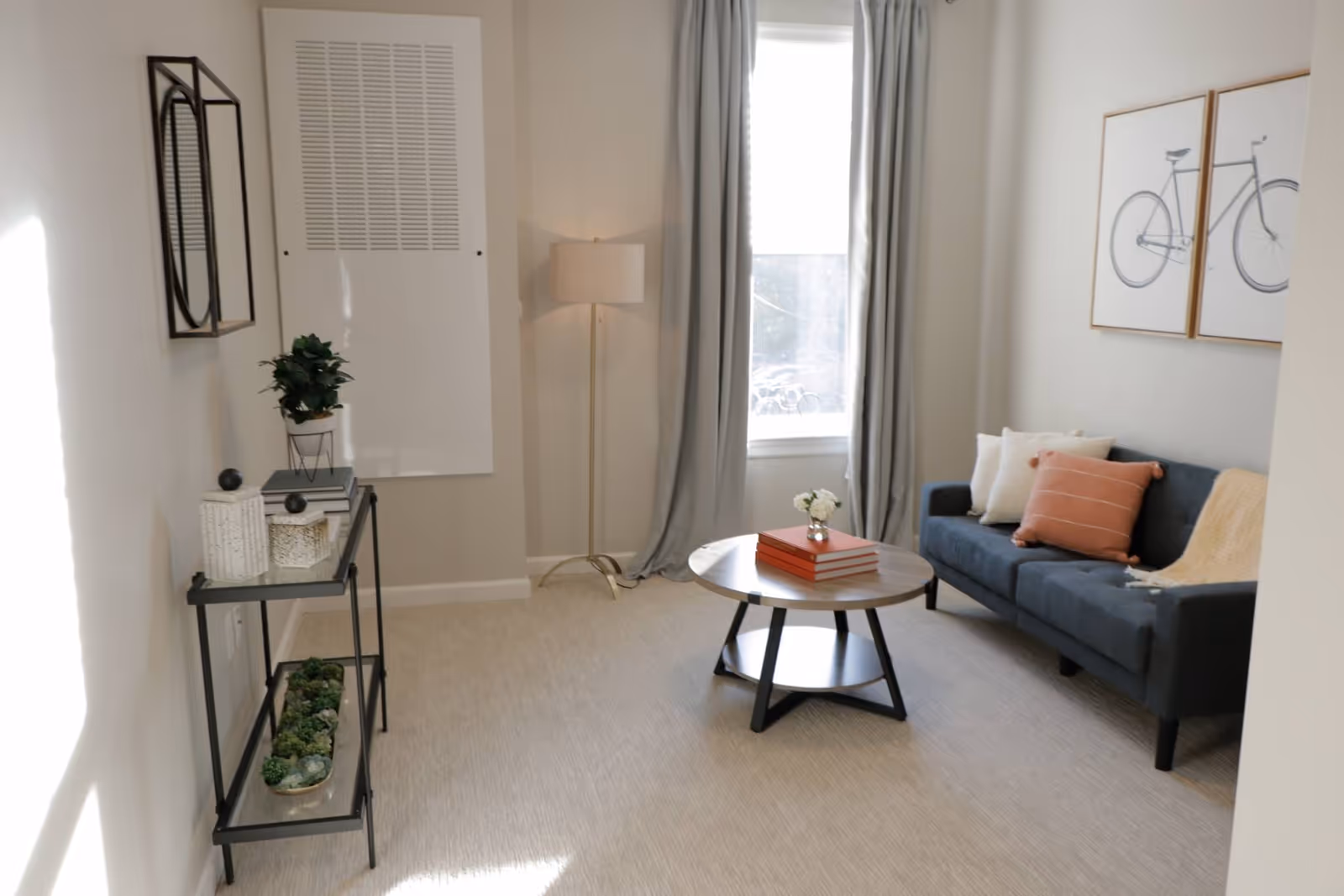 A cozy living room with a dark blue sofa adorned with white and orange pillows and a beige throw blanket. A round wooden coffee table with a small flower vase and stacked books sits in front of the sofa. Light gray curtains frame a window letting in natural light. On the left wall, there is a black metal console table with decorative plants and books, and a modern wall-mounted mirror above it. A tall floor lamp stands in the corner near the window. The walls are painted light gray, and the floor is covered with a light-colored carpet.