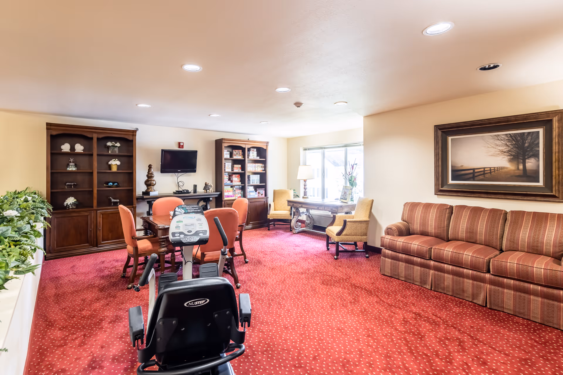 A senior living facility common room with red carpet, a striped sofa, two yellow armchairs, a wooden table with a lamp, two wooden bookshelves filled with items, a wall-mounted TV, a small table with four orange chairs, and an exercise bike in the foreground.
