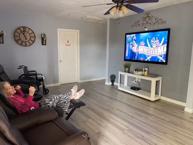 An elderly woman reclining on a brown leather chair with her feet up, watching a WrestleMania event on a wall-mounted flat screen TV in a spacious room with light gray walls and wood flooring. There is a white door with the number 6, a wheelchair, a small white console table with plants and items, a ceiling fan with lights, and a large wall clock.
