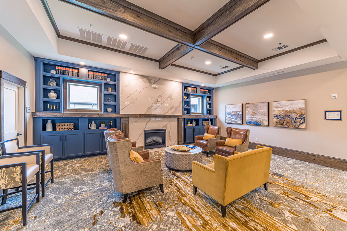 A cozy senior living facility common area with a modern fireplace centered on a marble wall. Surrounding the fireplace are built-in blue shelves with decorative items and books. The room features a patterned carpet in shades of gray and gold, several comfortable armchairs and a yellow loveseat arranged around a round coffee table. The ceiling has wooden beams and recessed lighting, and three abstract paintings hang on the beige wall.
