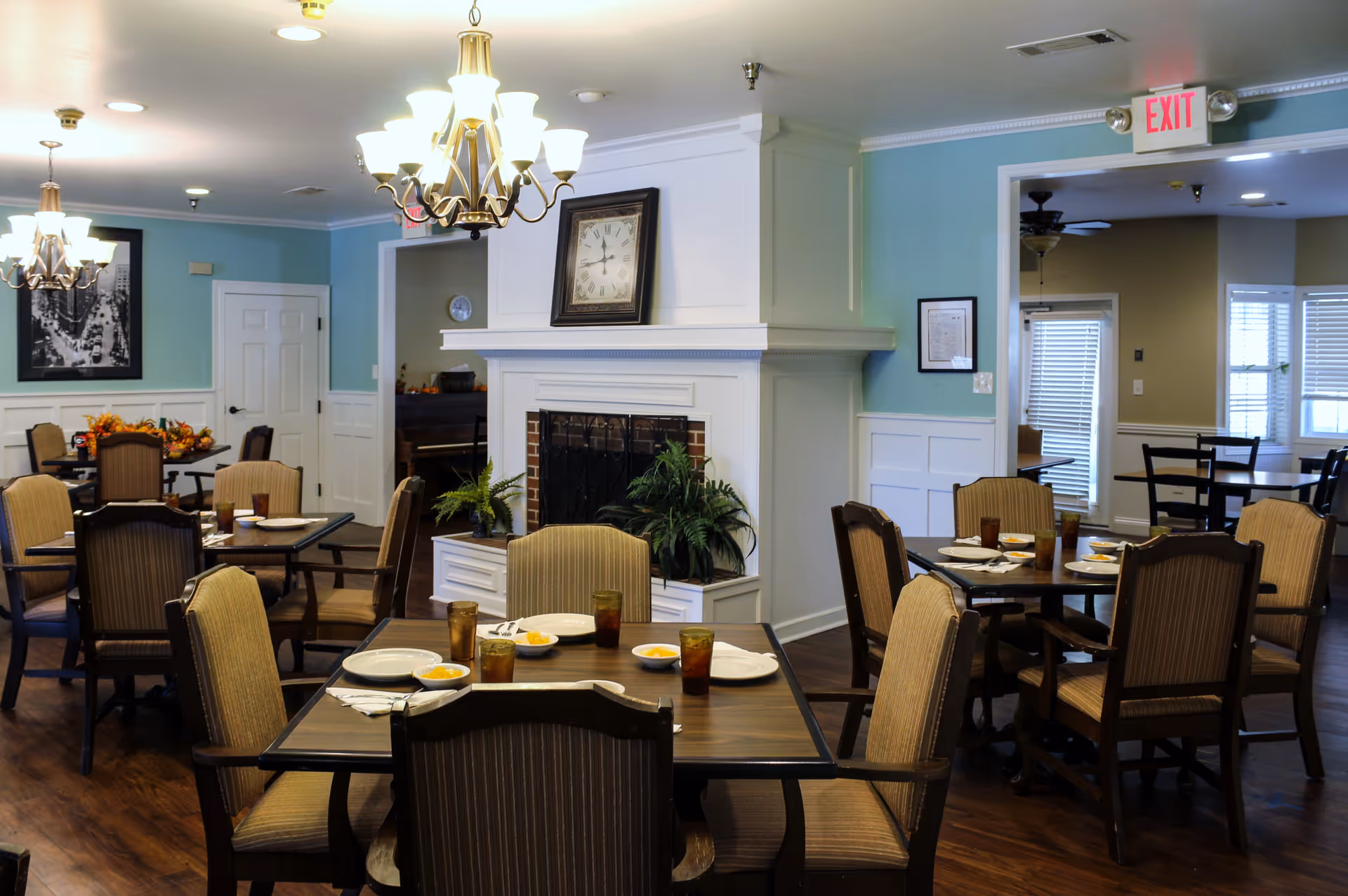 A dining room in a senior living facility with several tables set with plates, bowls of fruit, and glasses. The room features wooden floors, upholstered chairs, a white fireplace with a clock above it, and blue walls with white wainscoting. There are chandeliers hanging from the ceiling and an exit sign above a doorway.