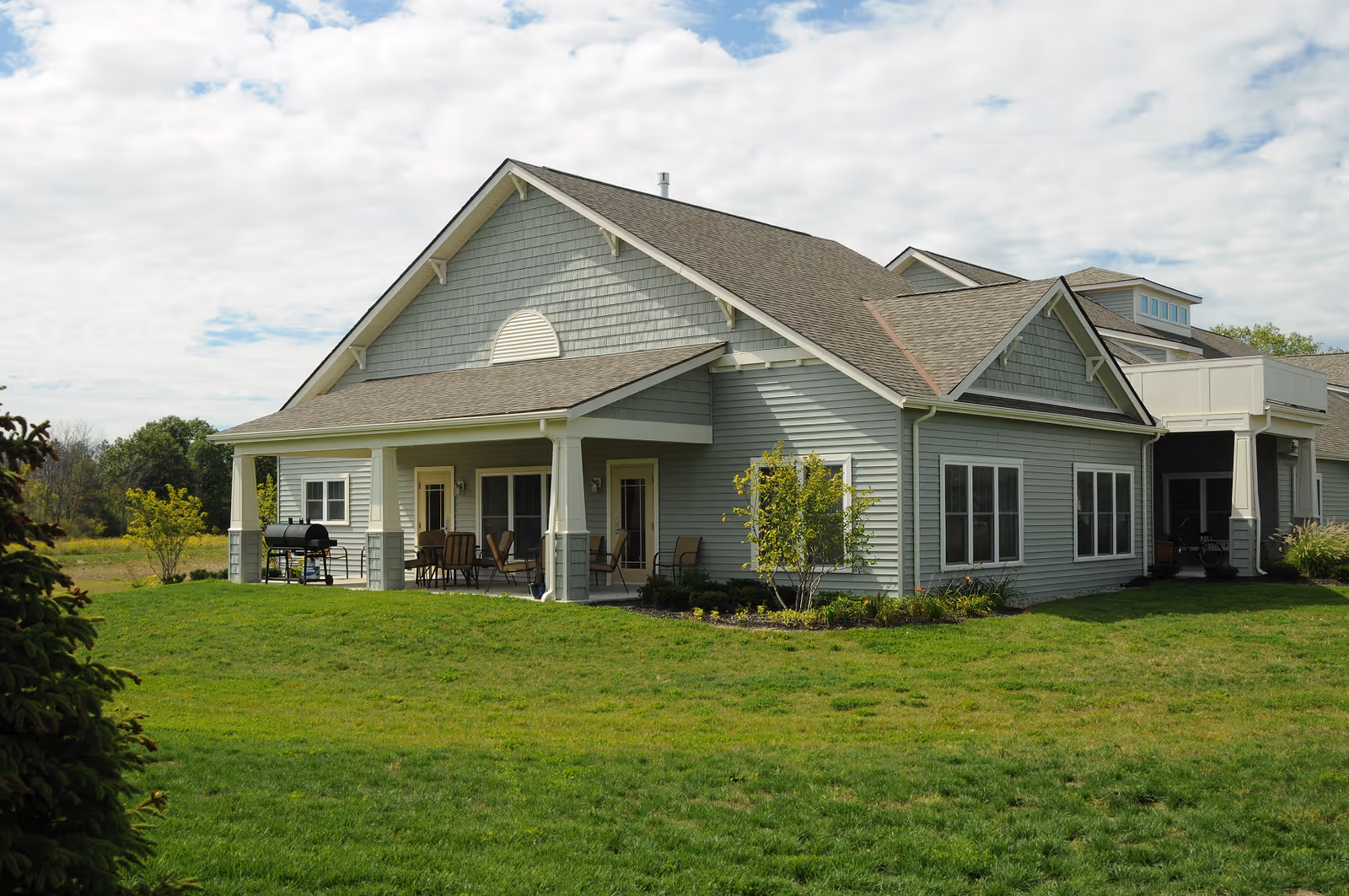 Gray single-story building with a covered front porch, outdoor seating and a grill on a grassy lawn under a partly cloudy sky.