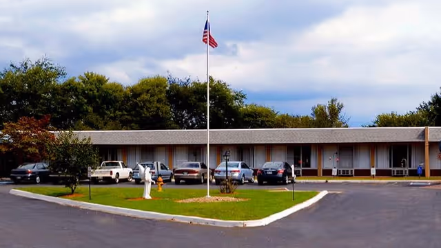 Single-story facility front with a circular driveway, parked cars, a flagpole flying an American flag, and lawn in front.
