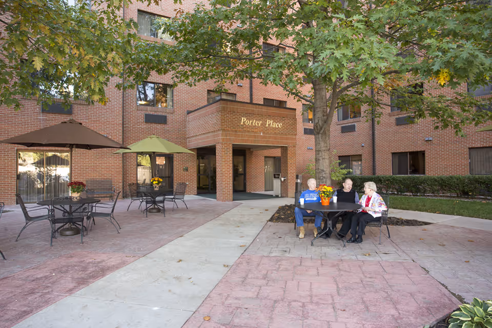 Outdoor patio area at Porter Place Retirement Community with three elderly people sitting at a round table under a tree. The patio has several tables with umbrellas and chairs, and the brick building entrance is visible in the background.