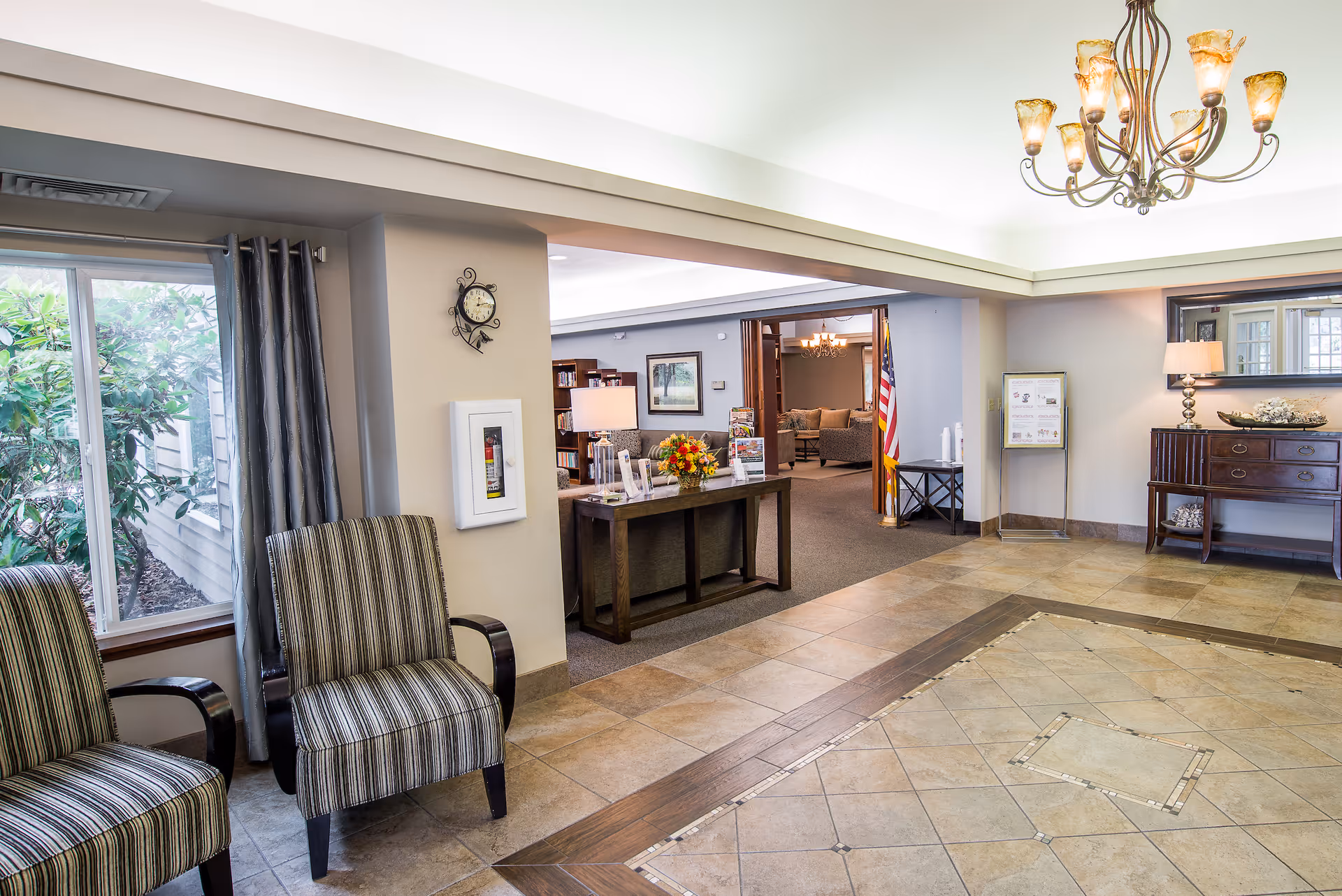 A bright and welcoming interior lobby area of a senior living facility with tiled flooring, two striped armchairs near a window with curtains, a wooden table with flowers and brochures, a wall clock, and a chandelier hanging from the ceiling. In the background, there is a seating area with sofas, an American flag, and a wooden sideboard with a lamp and decorative items.