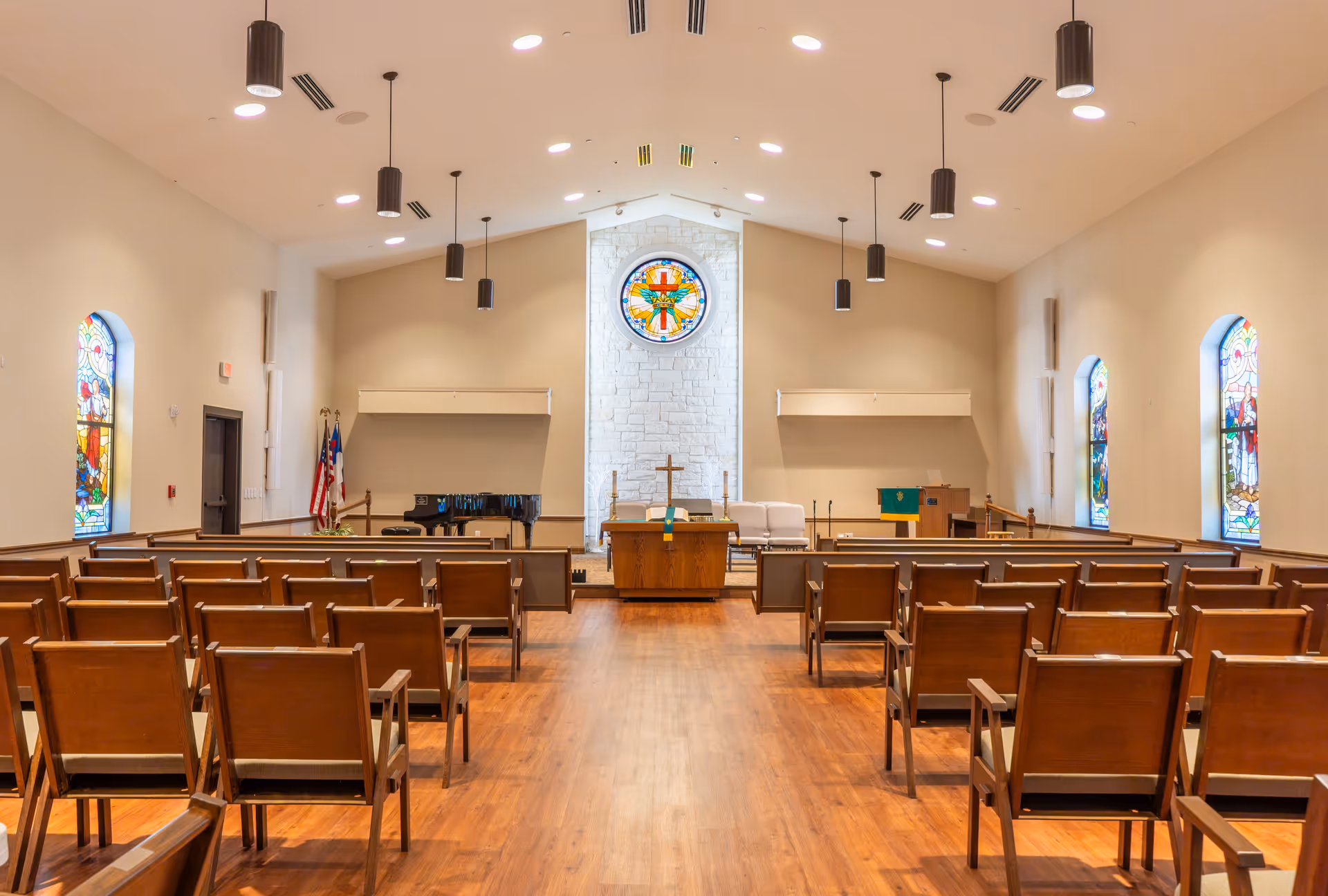 A chapel interior with rows of wooden chairs facing an altar, a cross, and stained glass windows including a circular window above the altar.
