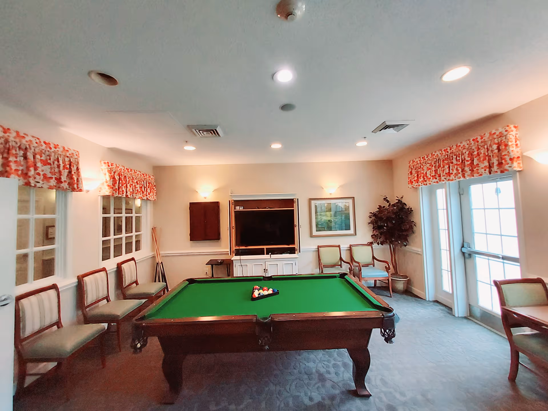 Interior room with a pool table in the center, surrounded by chairs along the walls. The room has windows with red and white patterned valances, a TV mounted on a cabinet, framed artwork, and a potted plant near a glass door.
