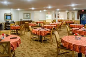 A spacious dining room with multiple round tables covered in red patterned tablecloths, each surrounded by wooden chairs. The room is well-lit with recessed ceiling lights, and there are framed pictures and a mirror on the walls. The carpet is beige with a subtle pattern.