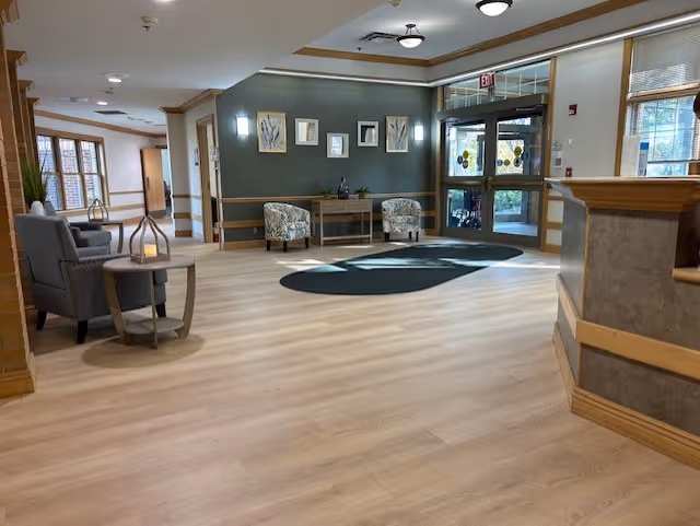 A spacious and well-lit interior common area with light wood flooring, a seating area with two patterned armchairs and a small table against a gray accent wall, and a glass exit door with an exit sign above it. There are additional chairs and a small table on the left side near windows, and a reception desk on the right side.