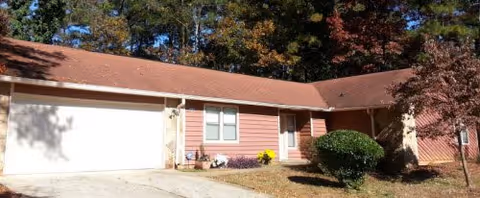 Single-story residential building with a brown shingled roof and light brown siding, featuring a two-car garage on the left, a front door with a small porch in the center, and a bush and small tree in the front yard under a clear sky.