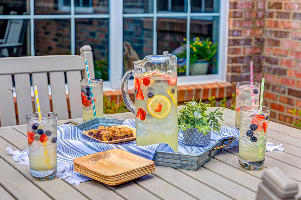 Outdoor patio table with a pitcher of fruit-infused lemonade and four glasses filled with lemonade and fresh berries, each with a colorful striped straw. The table also has a small potted plant, a tray with dessert bars, and wooden plates. The background shows a brick wall and a window with plants inside.