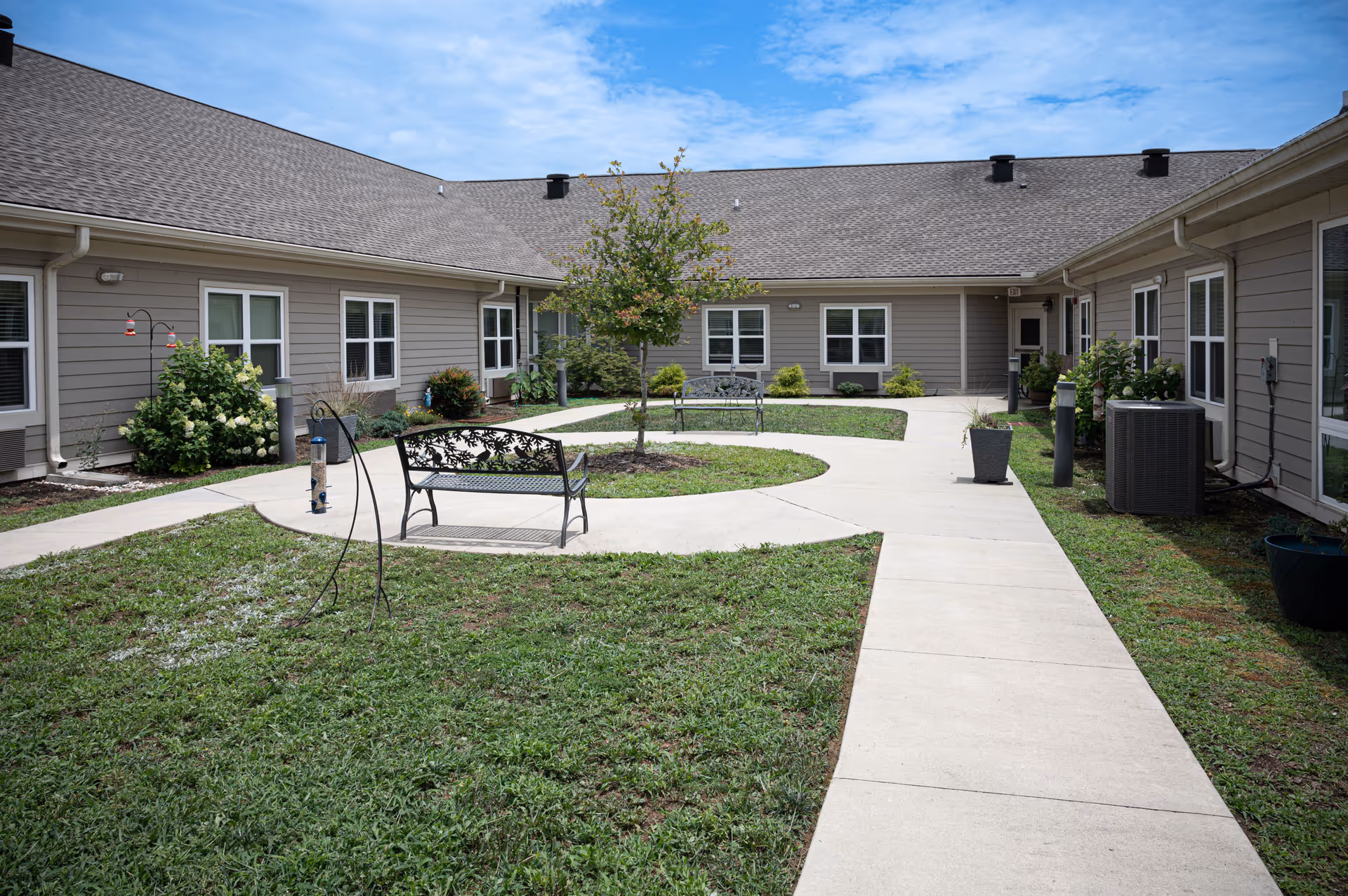 Outdoor courtyard area at The Landings of Mills River featuring a concrete walkway, green grass, a small tree in the center, two decorative metal benches, potted plants, and beige single-story building walls with multiple windows under a partly cloudy blue sky.