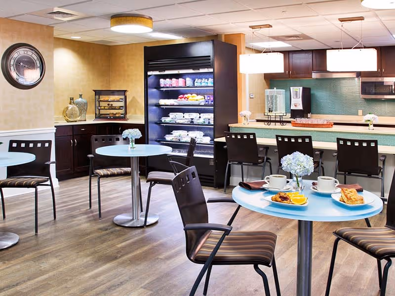 A bright and clean dining area in a senior living facility with round tables and chairs. One table in the foreground has plates with breakfast food, coffee cups, and a small vase with blue flowers. In the background, there is a refrigerated display case with drinks and food items, a counter with bar stools, and kitchen appliances including a microwave and coffee machine.