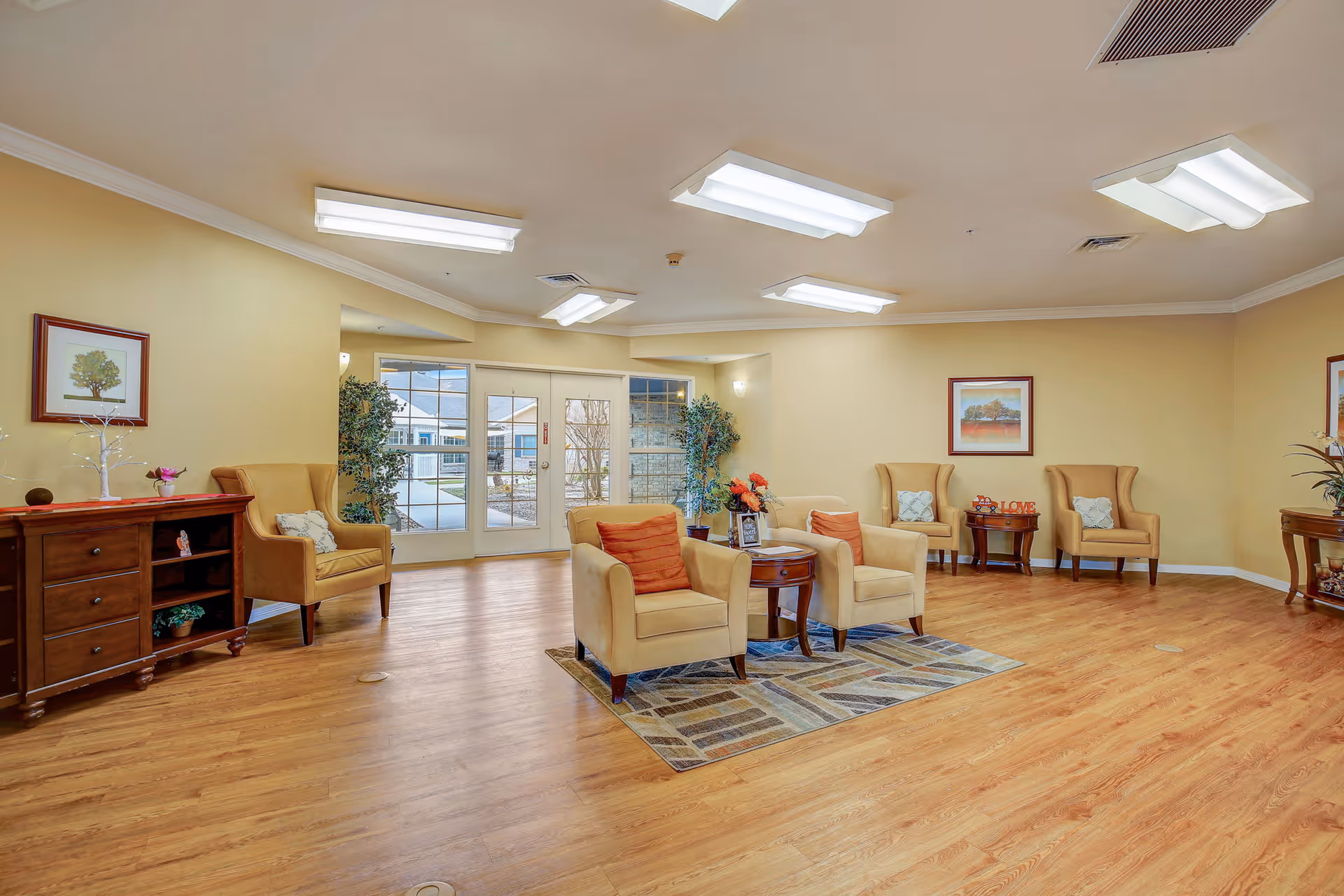 A spacious and well-lit living room area with wooden flooring, beige walls, and multiple beige armchairs arranged around small wooden tables. The room features framed artwork on the walls, potted plants, and large glass doors leading to an outdoor area.