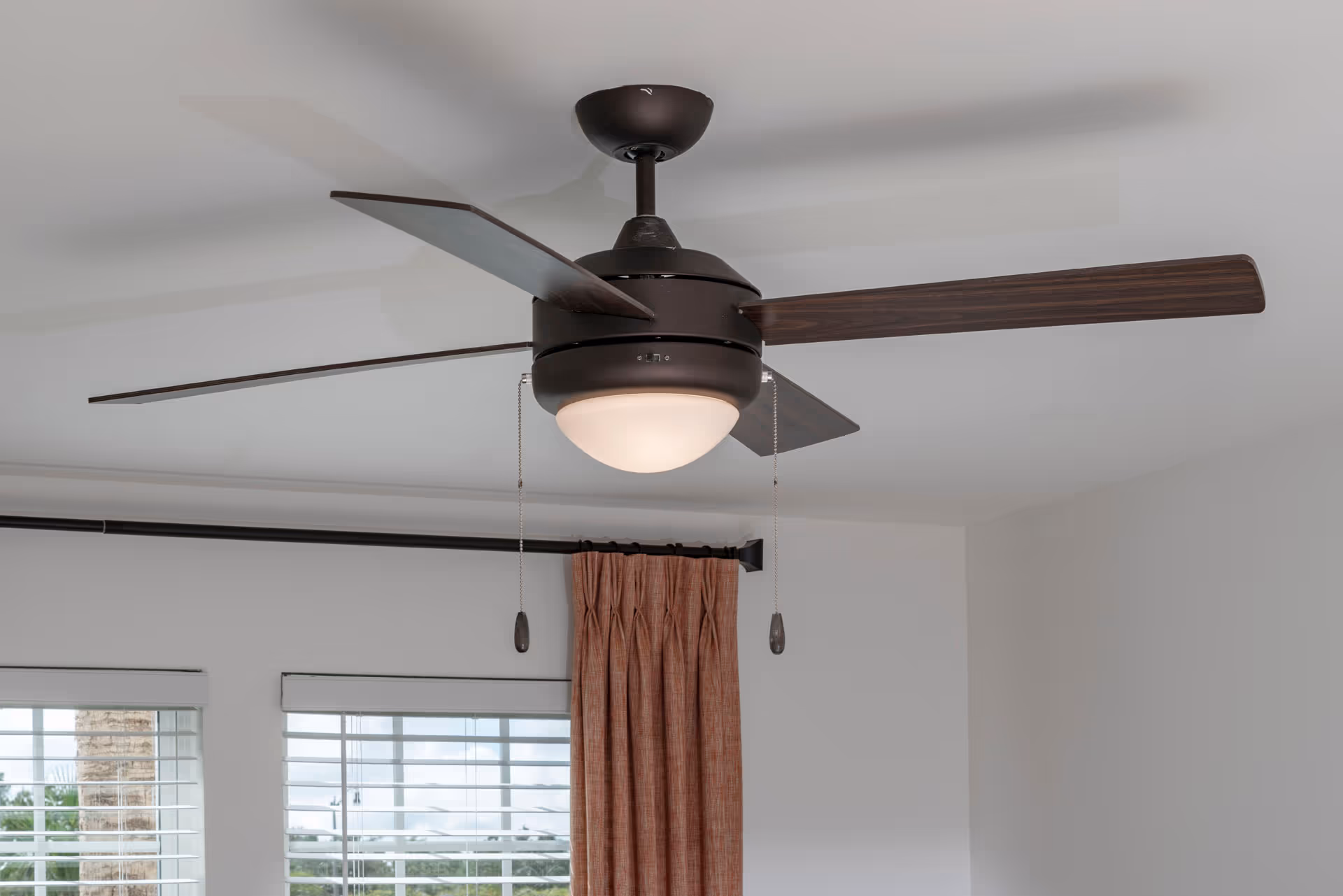 A modern ceiling fan with dark wooden blades and a central light fixture mounted on a white ceiling. Below the fan, there are windows with white blinds and a curtain rod with rust-colored curtains.