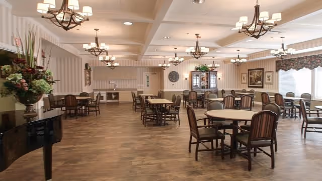 A spacious dining room with multiple wooden tables and chairs arranged neatly. The room features elegant chandeliers hanging from a coffered ceiling, striped wallpaper, large windows with valances allowing natural light, and decorative floral arrangements. A piano is visible on the left side of the room.