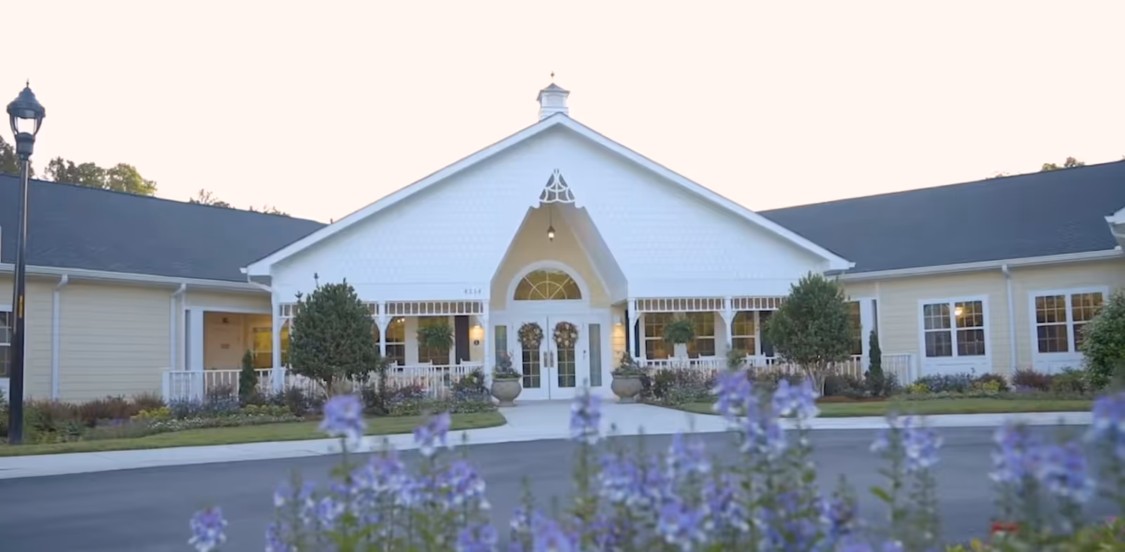 Front exterior view of a single-story building with a peaked roof and white double doors decorated with wreaths. The building is surrounded by landscaping including bushes and purple flowers in the foreground, with a lamppost on the left side.