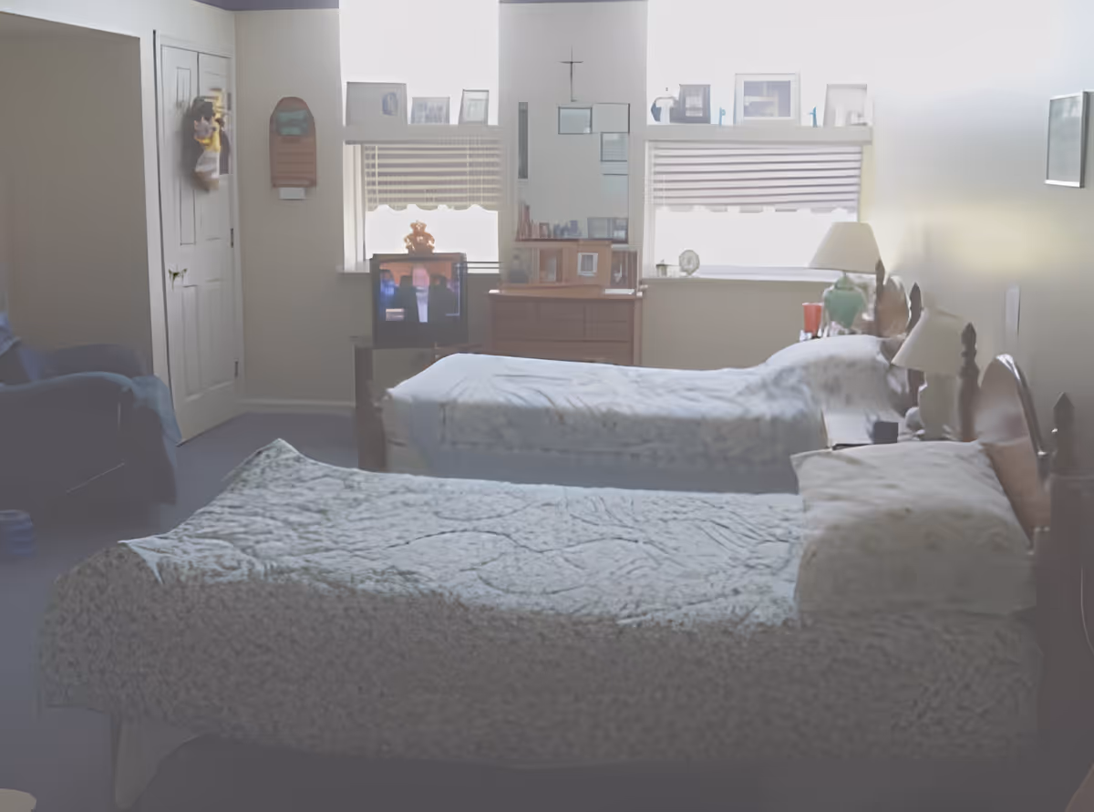 Two twin beds in a softly lit bedroom with a dresser, small TV, lamp, and an armchair.