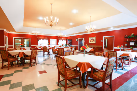 A spacious dining room with multiple tables covered with white and orange tablecloths, each adorned with a small vase of flowers. The room has red walls, chandeliers hanging from the ceiling, and patterned chairs around the tables. There are windows with curtains and a clock on the wall.