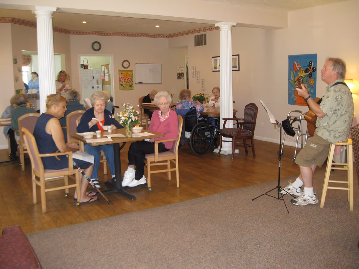 A group of elderly people sitting at tables in a communal room, some eating and others socializing. A man is seated on a stool playing a guitar near a music stand. The room has wooden floors, white columns, and light-colored walls with decorations and a clock.