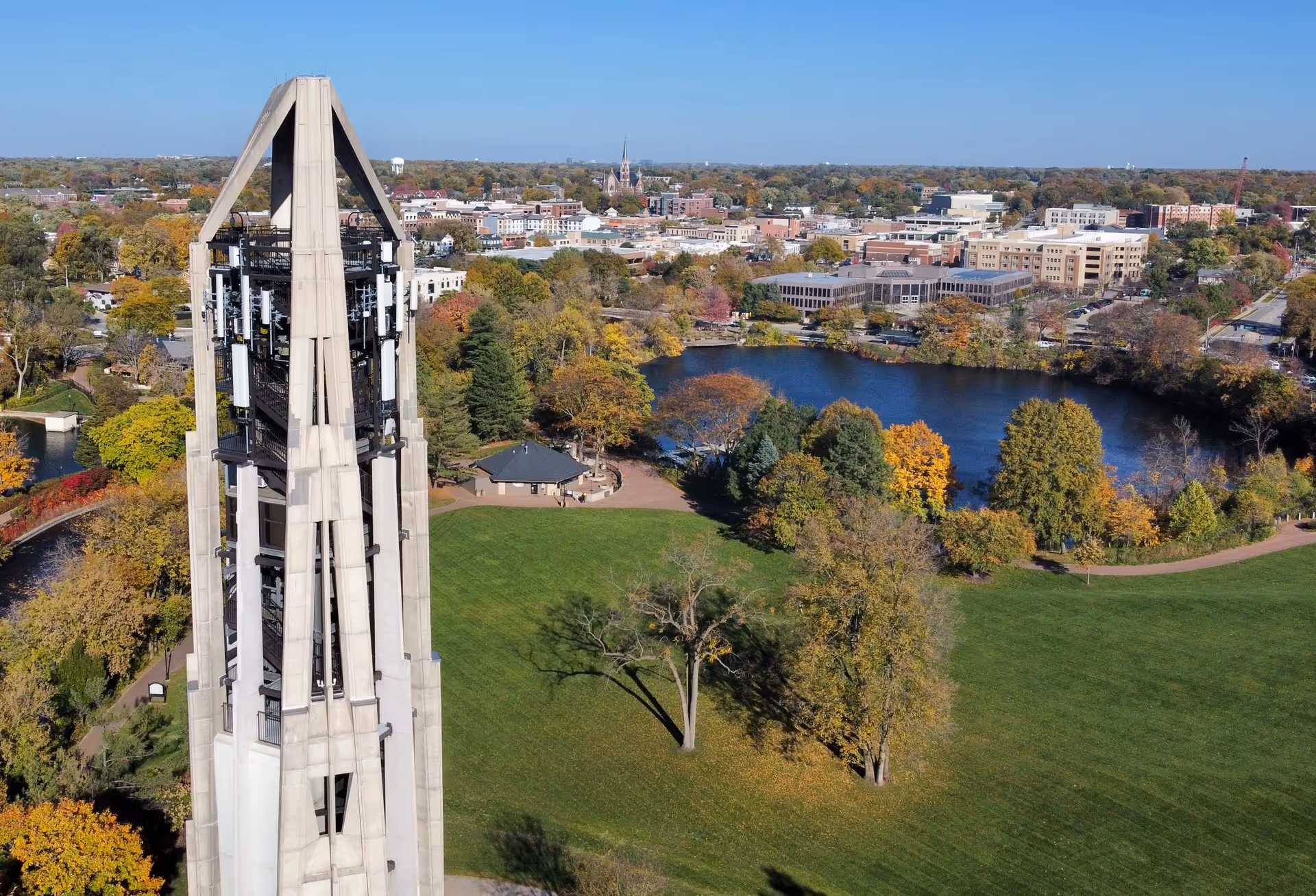 Aerial view of a tall concrete carillon tower beside a green park and pond with a city skyline in the background.