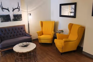 A cozy sitting area with a dark gray tufted sofa, two bright yellow wingback chairs, a round marble-top coffee table with a decorative black wire bowl, a floor lamp, and wall art featuring abstract antlers. A rectangular mirror hangs on the wall above one of the chairs.