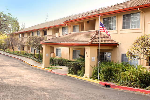 Two-story beige senior living building with red-tiled roof and American flag at entrance.