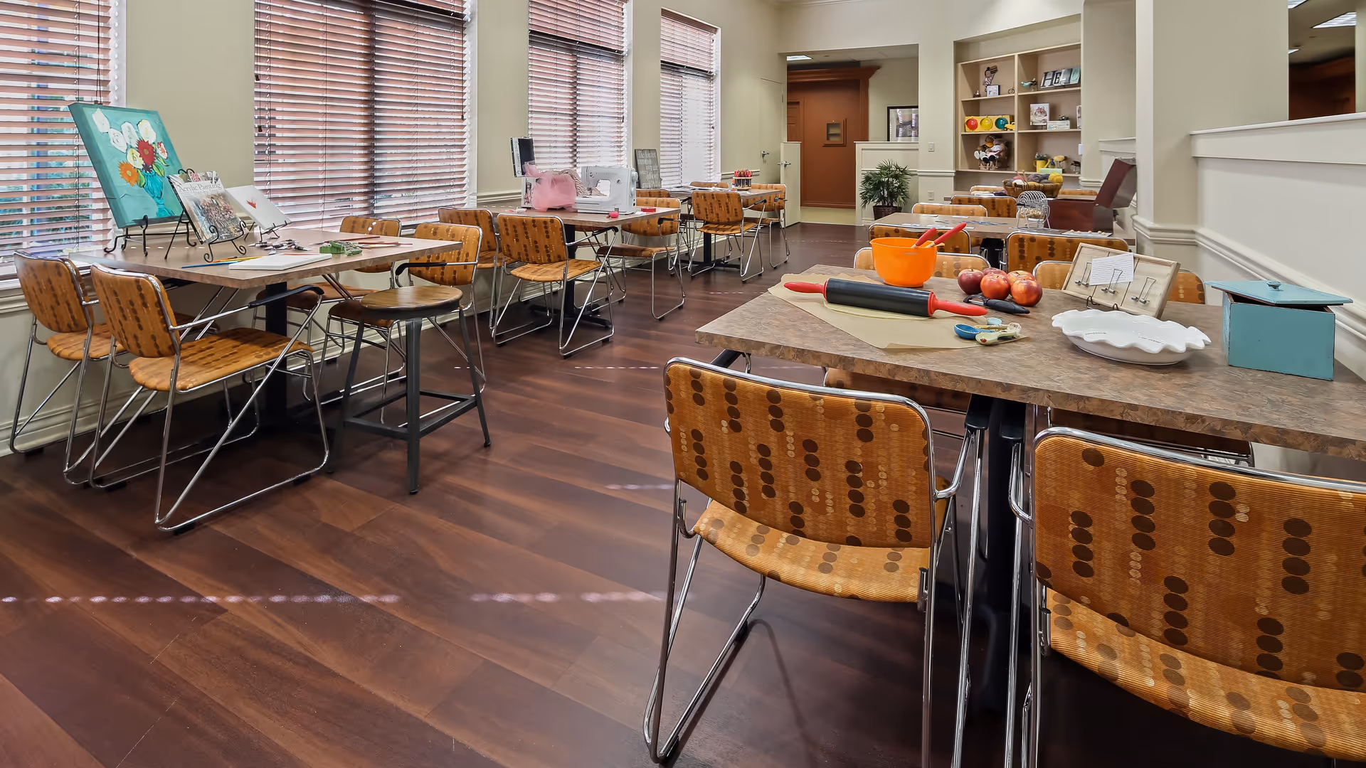 A well-lit activity room with multiple tables and chairs arranged for group activities. The tables have various craft supplies including a rolling pin, apples, a bowl, scissors, and paper. Large windows with wooden blinds allow natural light to fill the room. Shelves in the background hold decorative items and supplies.