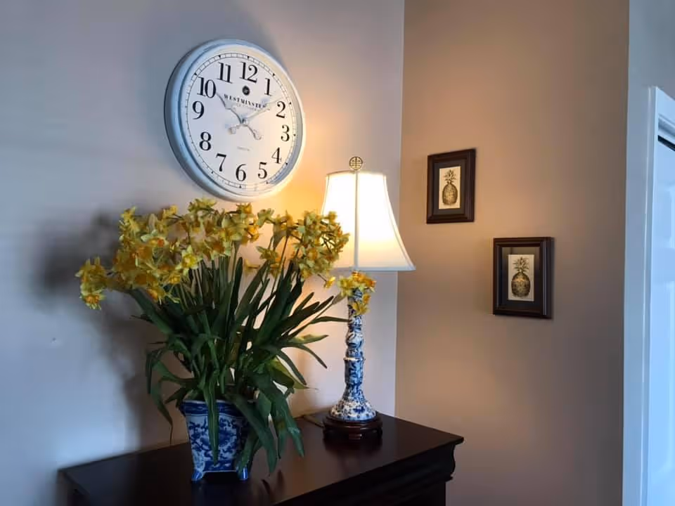 A corner of a room with a dark wooden table holding a blue and white vase filled with yellow flowers and a matching blue and white table lamp with a white lampshade. Above the table is a large round wall clock showing the time as 10:10. On the adjacent wall, there are two framed pictures of pineapples.